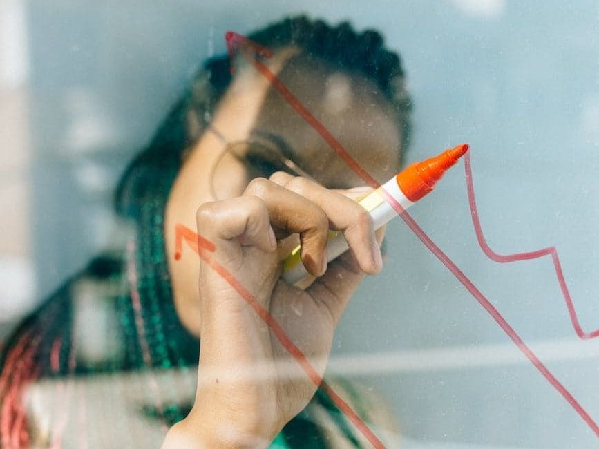 woman in a beige coat writing on a glass panel using a whiteboard marker